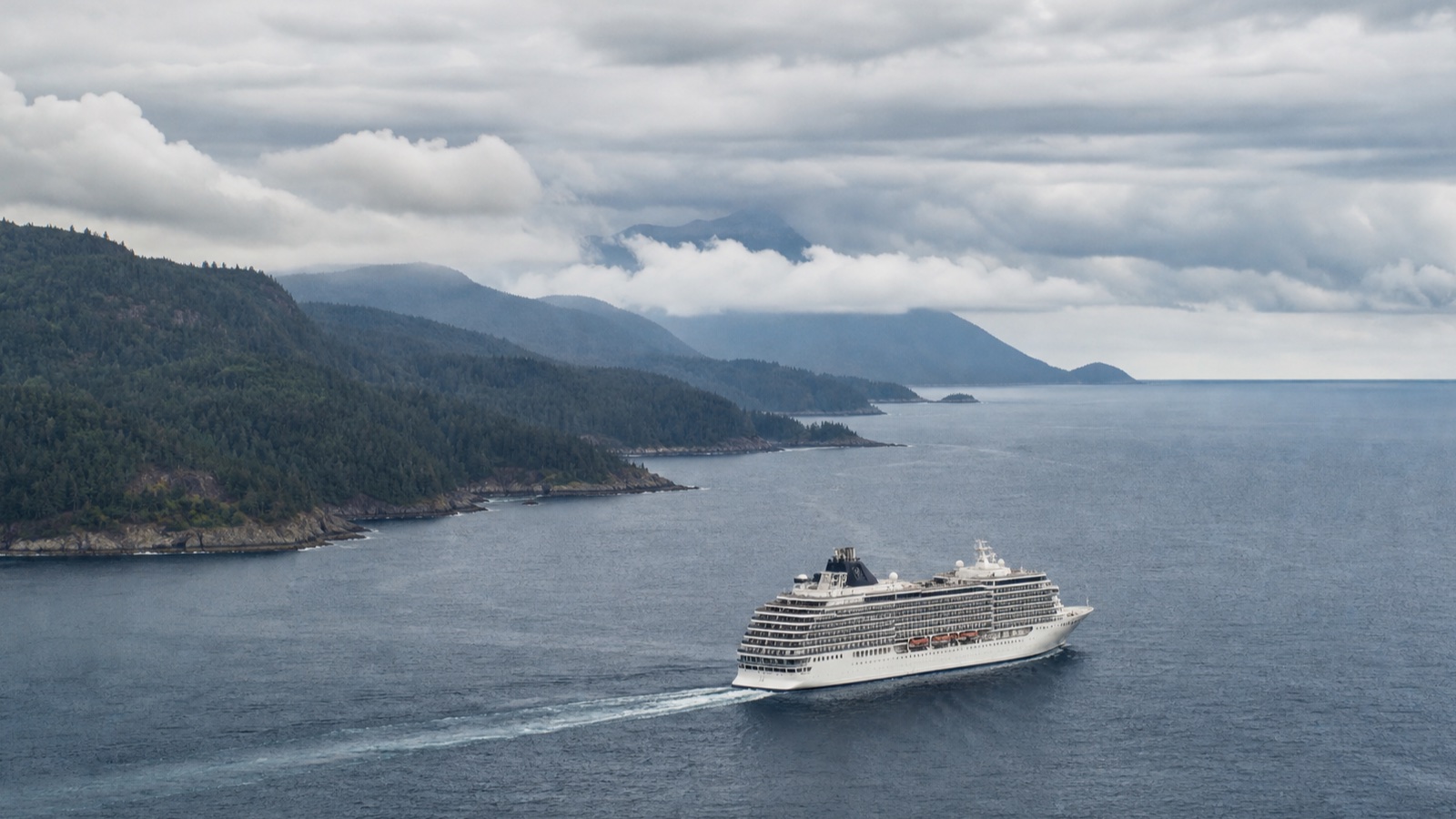 Unbranded cruise ship sailing past a forested Pacific Coast under changeable weather
