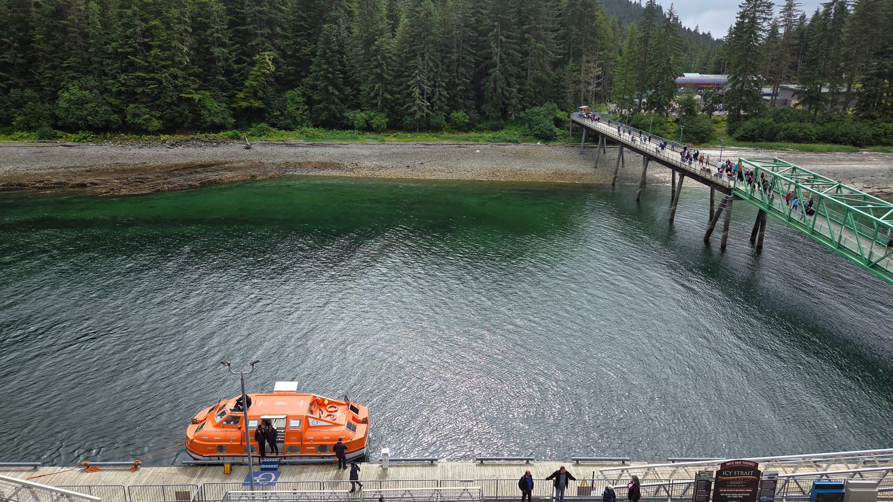 Green gangway crossing clear water between a cruise ship and the forested shore at Icy Strait Point